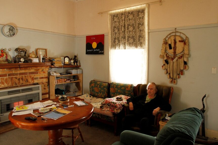 Shirley Wilson sits in an armchair in a country lounge room. Aboriginal artwork and a photo of Audrey Hepburn are on the wall.