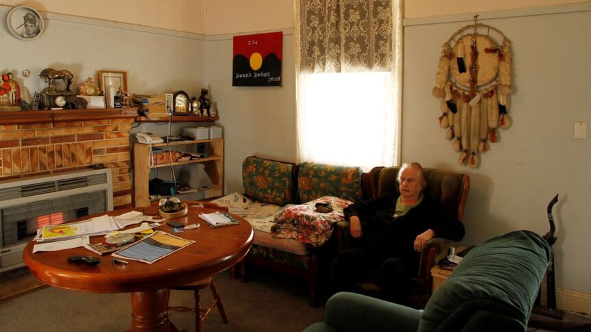 Shirley Wilson sits in an armchair in a country lounge room. Aboriginal artwork and a photo of Audrey Hepburn are on the wall.