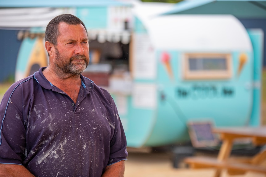 A man standing looking serious, with an out of focus food caravan in the background