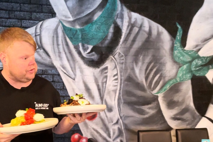 A man carrying two plates of food in front of a mural of a chef wearing an headband over his eyes