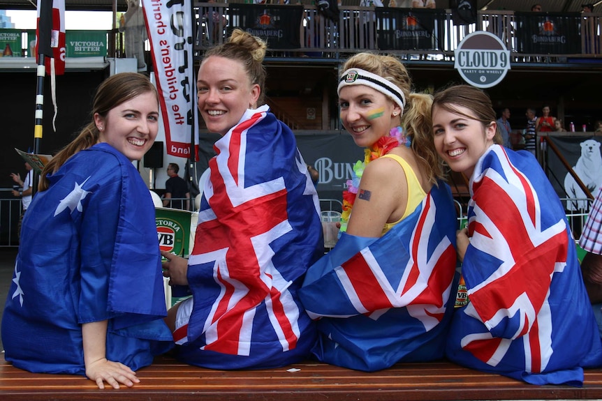 Four young women draped in Australian flags sit on a bench at a bar.