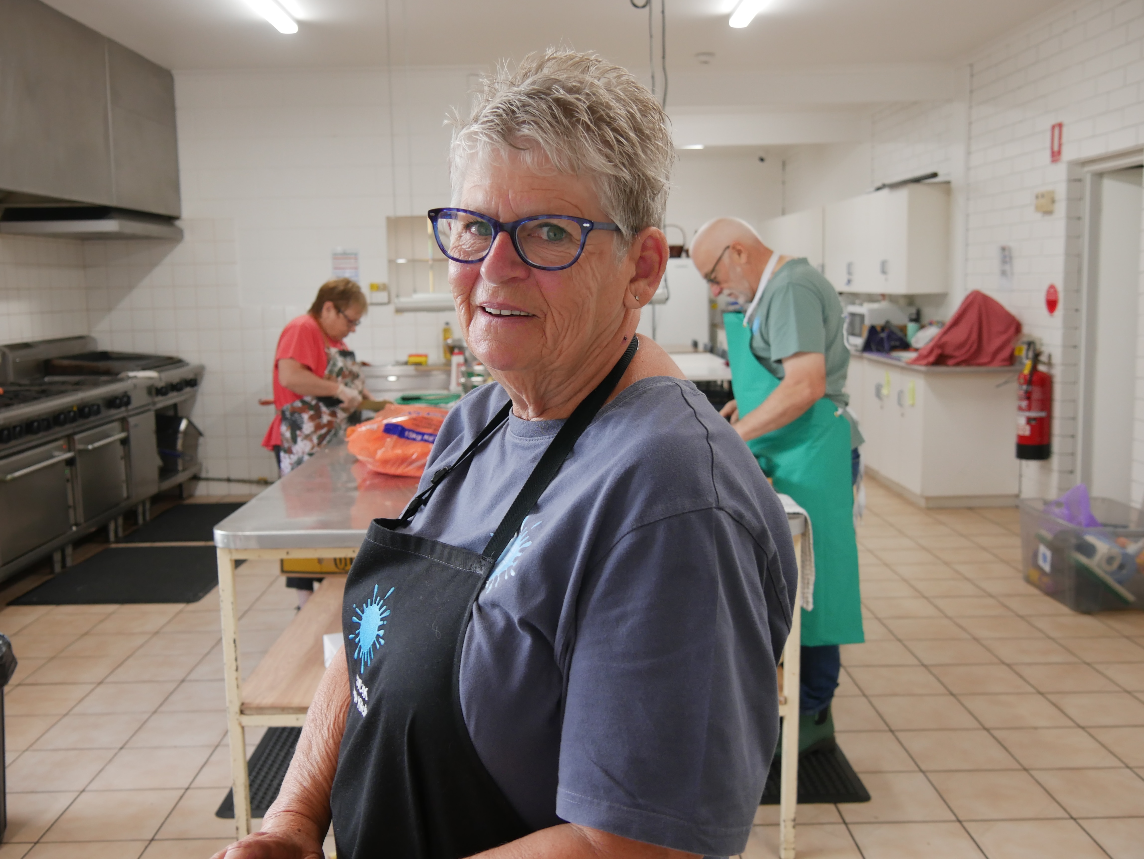A woman wearing an apron smiles at the camera while people work in a kitchen behind her. 