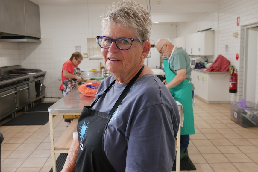 A woman wearing an apron smiles at the camera while people work in a kitchen behind her. 