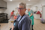A woman wearing an apron smiles at the camera while people work in a kitchen behind her. 
