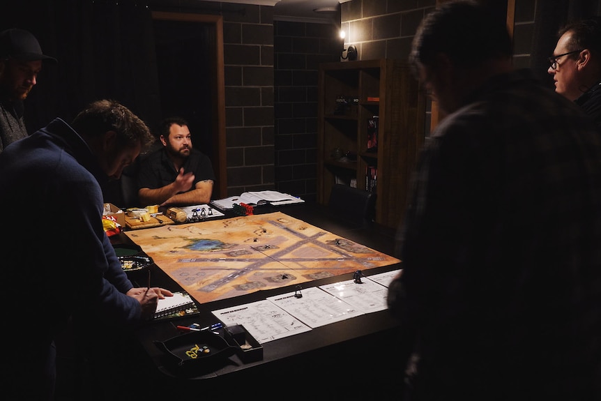 A group of six men are gathered around a sci fi table top game in a low-lit room at night.