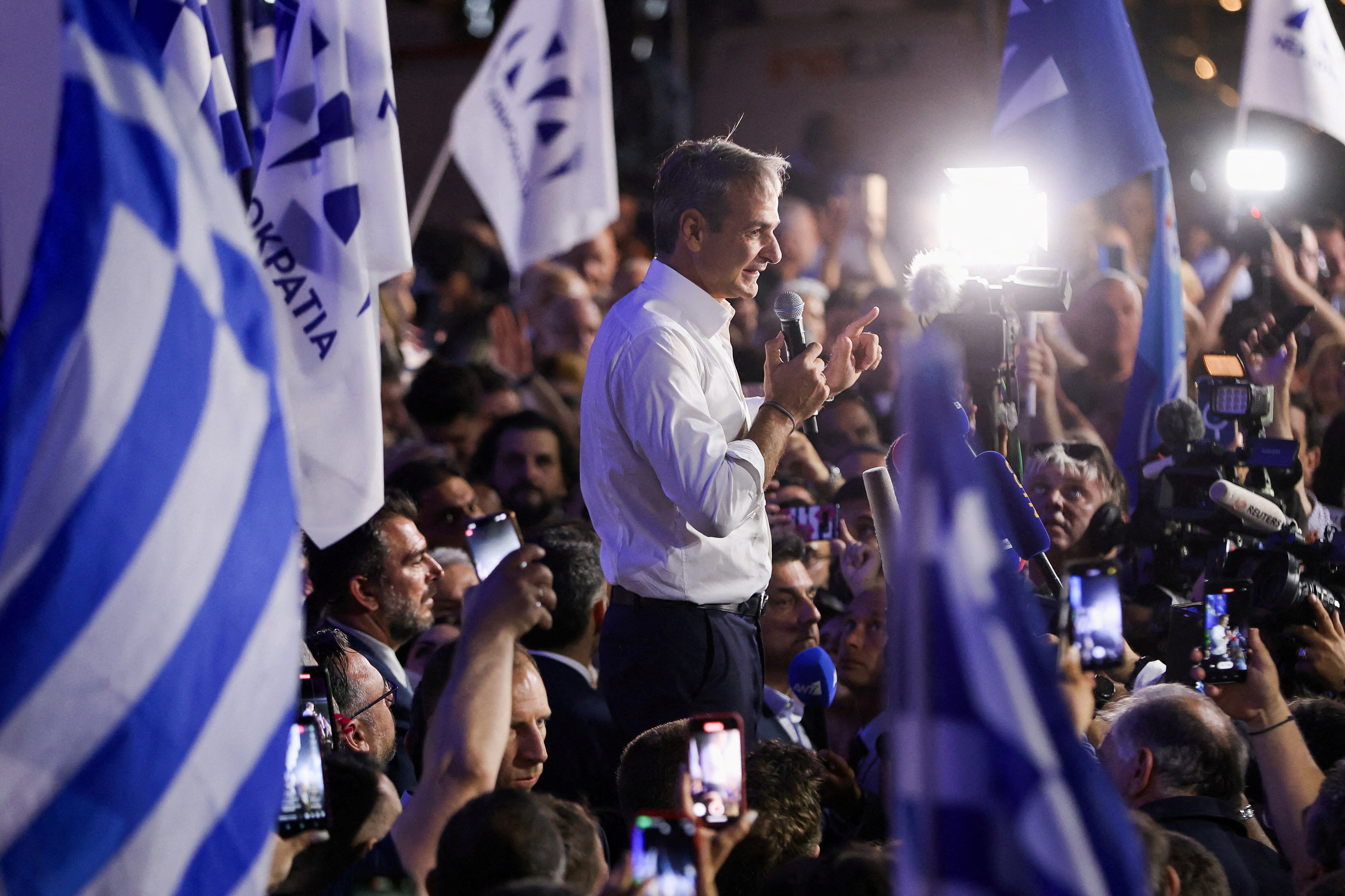 A Greek man in white shirt speaking to crowd amid blue and white Greek flags, cameras and lights.