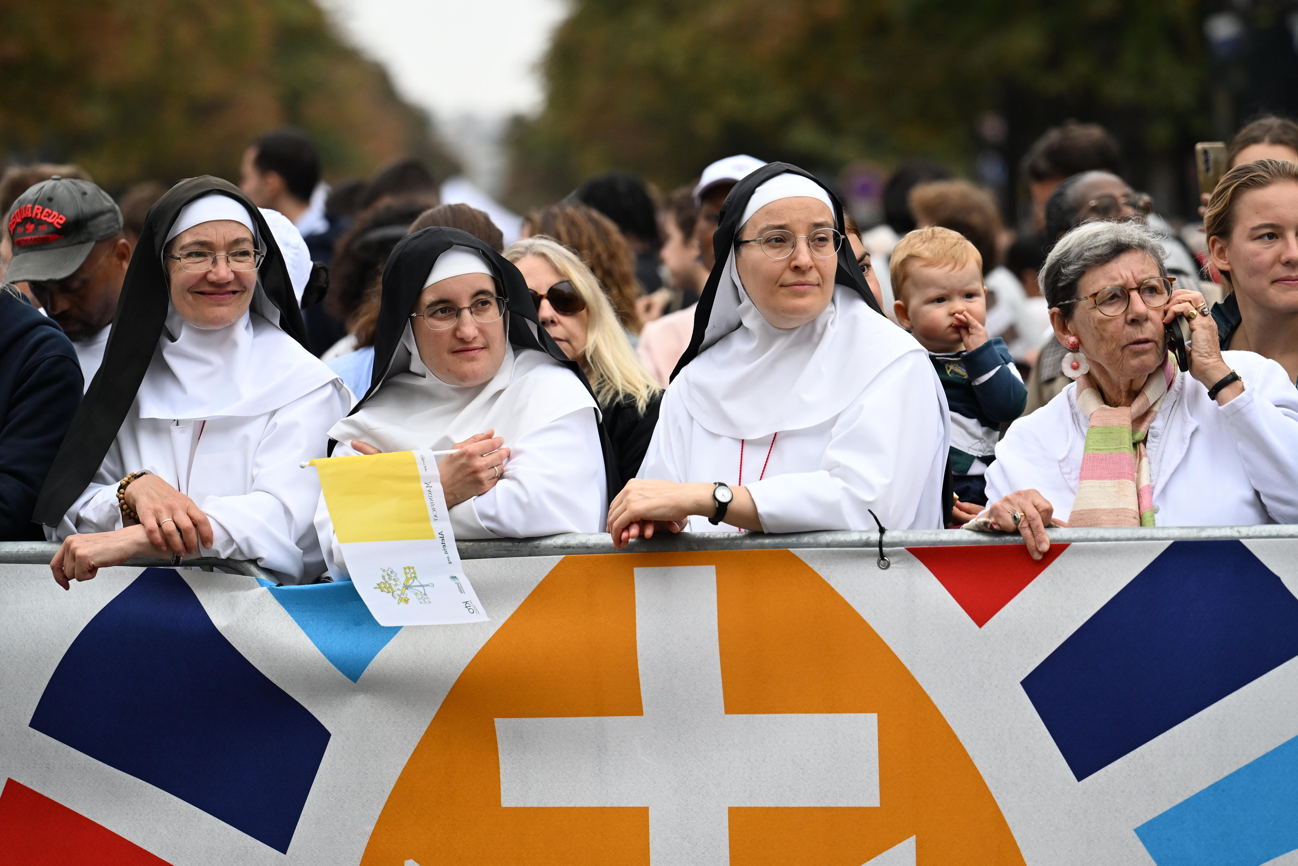 Three nuns stand behind a barricade, smiling and looking out, behind them is a large crowd of people.