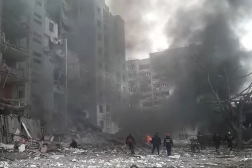Buildings filled with smoke are destroyed as people stand in rubble below.