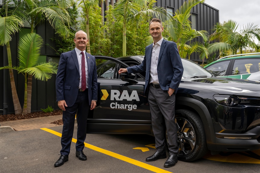 Two men in suits stand in front of car marked RAA charge