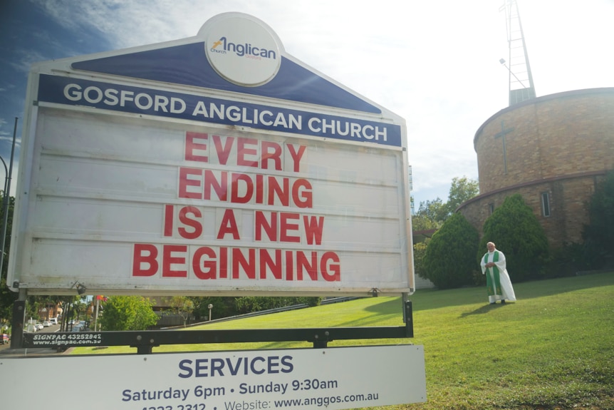 Priest in background and sign in foreground which reads Every ending is a new beginning 