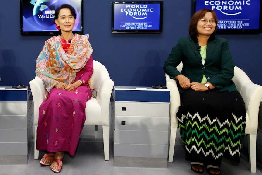 Burmese politicians sit in traditional outfits with screens in the background.