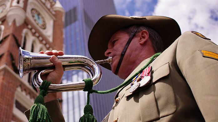 Upshot of a uniformed army bugler playing his bugle with the Perth Town Hall in the background.