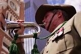 Upshot of a uniformed army bugler playing his bugle with the Perth Town Hall in the background.
