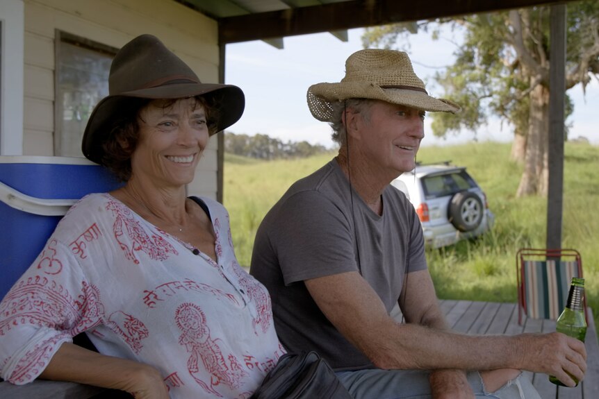 A couple sit on a veranda with a green hill and trees behind them.