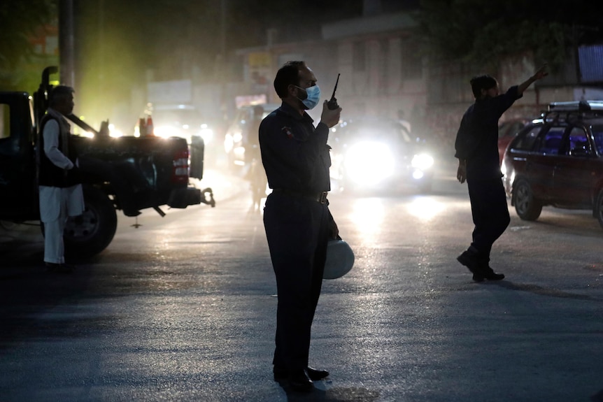 Afghan security personnel standing on a street at night holding a walkie-talkie, surrounded by patrol vehicles. 