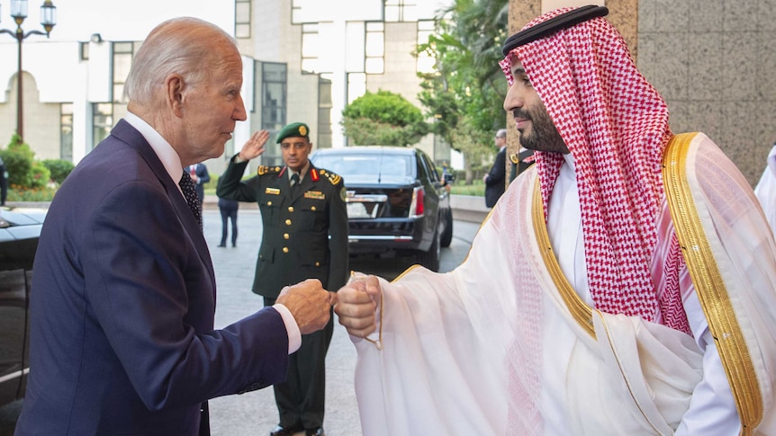 man in suit and man in traditional Arab dress, looking at each other and holding their bent knuckles together