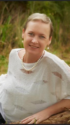 Woman with short, blonde hair, wearing a sheer, white top with cutouts over a white camisole, smiling as she poses for camera