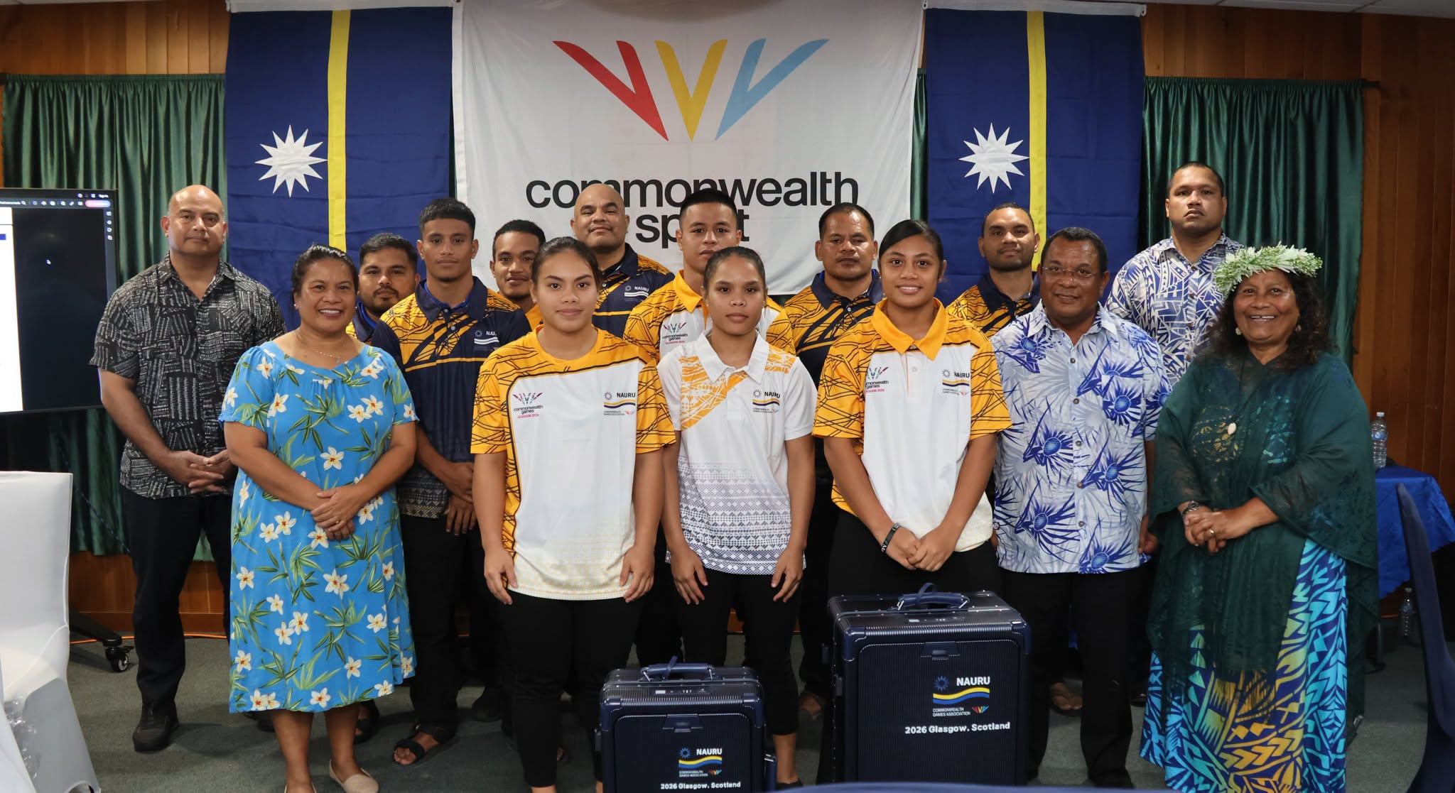 A group of athletes from Nauru wearing white and yellow athletics uniforms stand in a group with other people in a room smilling