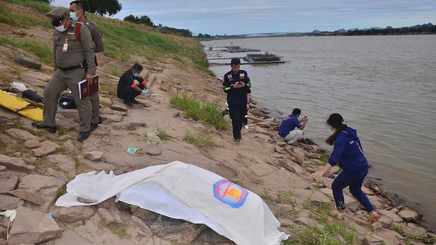 Thai rescuers cover a body on the shore of the Mekong River. Police officers with facemasks are standing nearby.