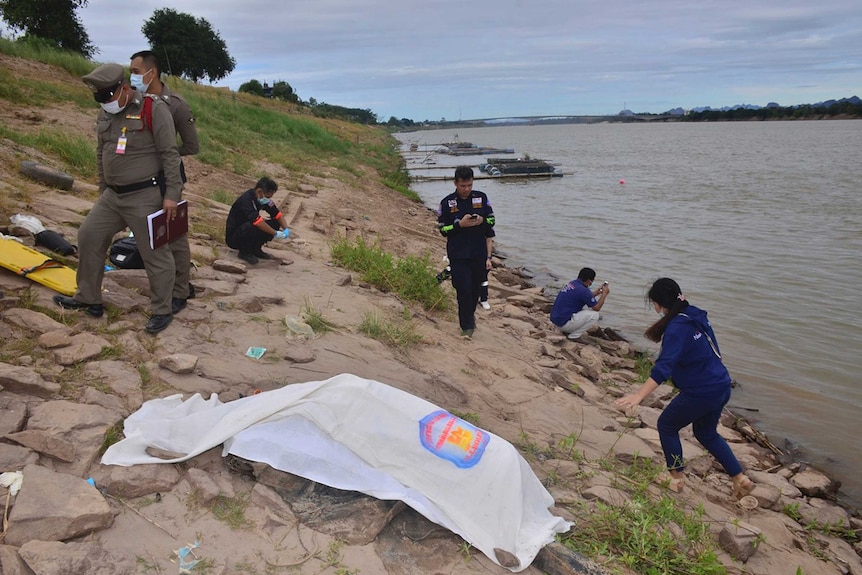 Thai rescuers cover a body on the shore of the Mekong River. Police officers with facemasks are standing nearby.