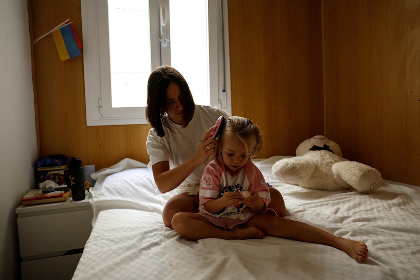 A woman brushes the hair of a girl sitting on a bed. 