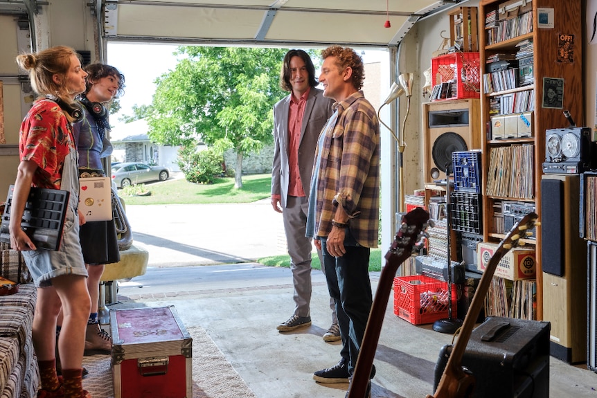 Two young women (one holding pad controller) stand facing two middle aged men in garage with guitar, amp, stacked vinyl and cds.