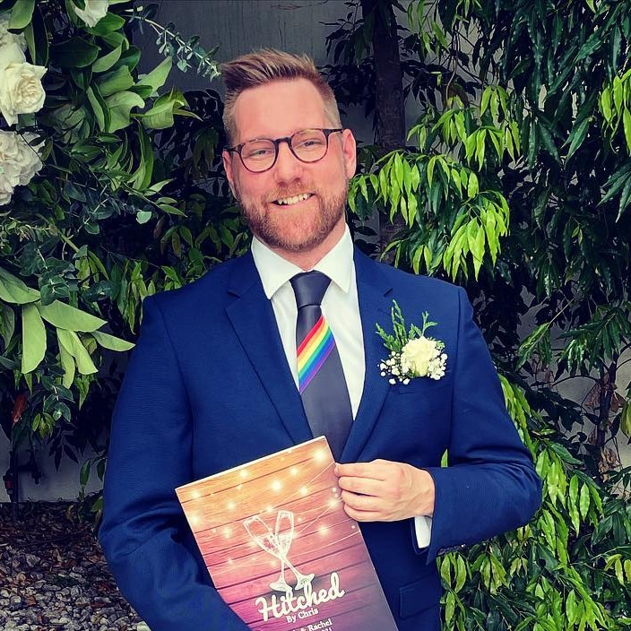 A smiling man in a blue suit stands in front of a flower arrangement