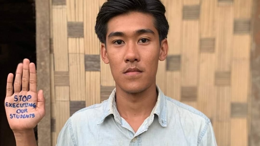 A Burmese man in collared shirt holds up hand with 'stop executing our students' on it