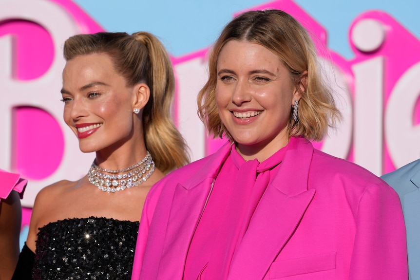A close up of Greta smiling at the Barbie premiere in a hot pink suit. Margot in next to her in a diamond necklace.