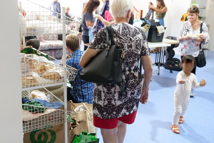 An older woman stands next to a shelf of bread while other people look at clothes and move around in the background.