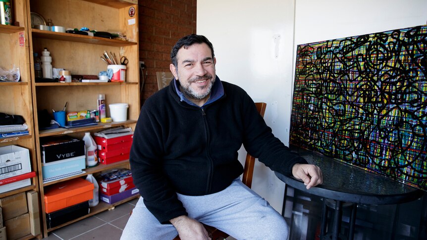 Artist in blue shirt, navy jumper and grey track pants sitting on stool in converted garage studio, smiling at camera.