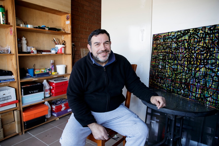 Artist in blue shirt, navy jumper and grey track pants sitting on stool in converted garage studio, smiling at camera.