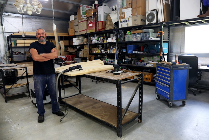 A man with short hair and beard stands with his hands crossed in a studio filled with wood-carving tools.