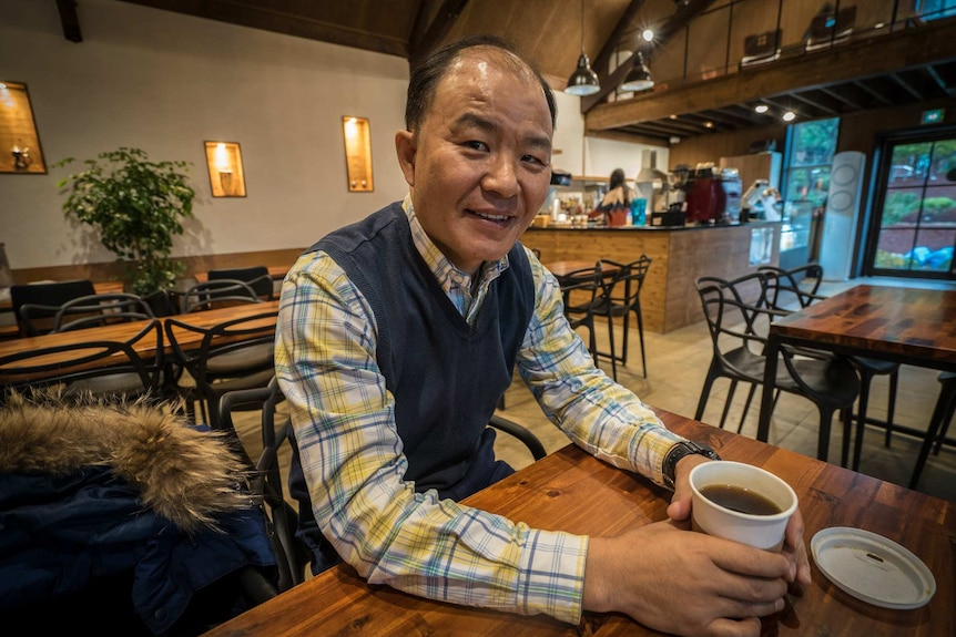 A middle-aged balding Asian man sits in a cafe nursing a black coffee in a paper cup with its lid laying upside down.