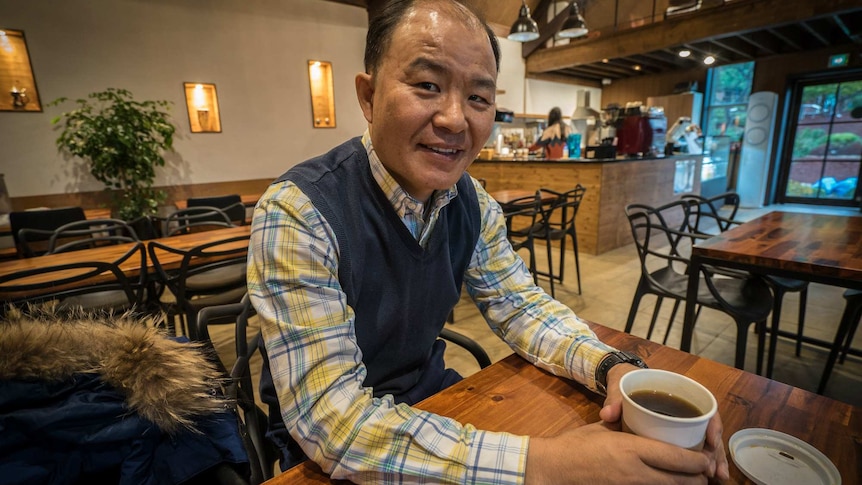 A middle-aged balding Asian man sits in a cafe nursing a black coffee in a paper cup with its lid laying upside down.