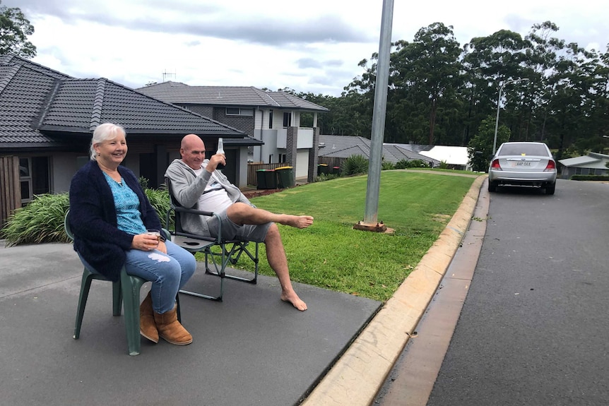 A man and a woman sitting on chairs on their driveway, holding drinks.