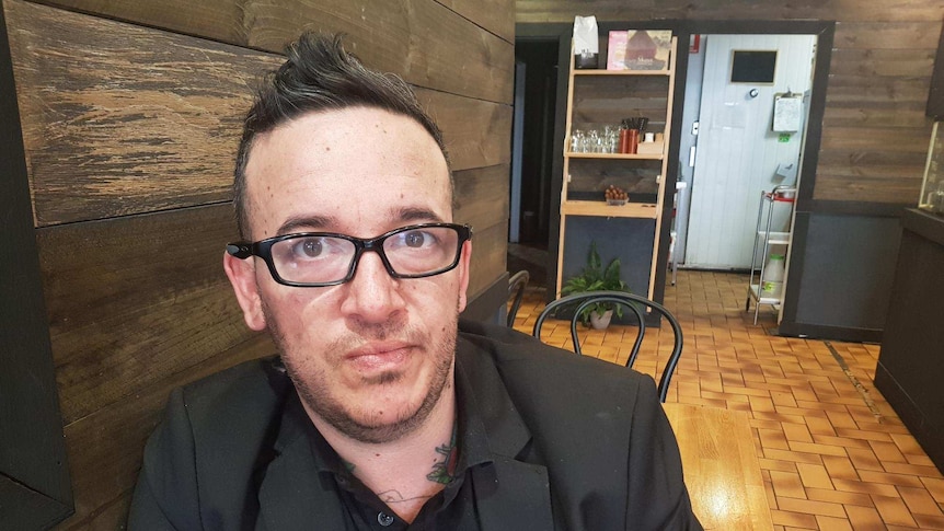 Close up of a man sitting in a cafe, wearing black shirt and jacket and glasses