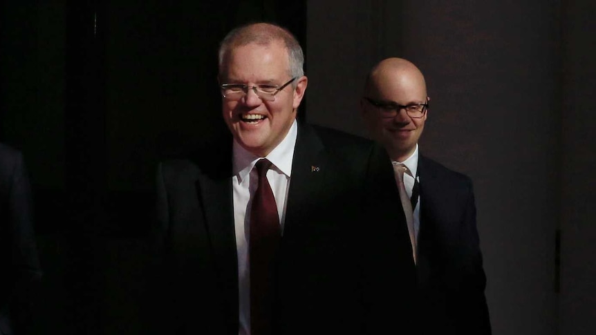 Scott Morrison smiles and laughs as he walks in shadows in Parliament house - two men stand behind him