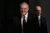 Scott Morrison smiles and laughs as he walks in shadows in Parliament house - two men stand behind him