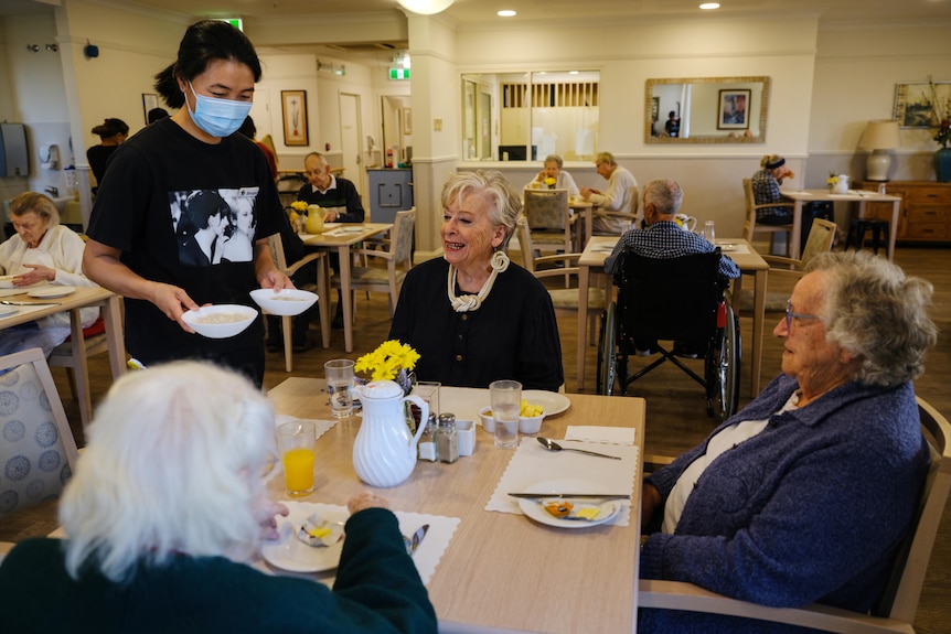 Maggie Beer and two female aged care residents being served porridge for breakfast in the dining hall.