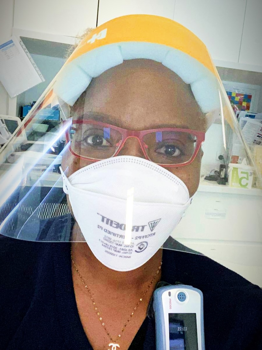 A nurse from Zimbabwe wearing a mask and face shield and pink glasses, holding a phone up.