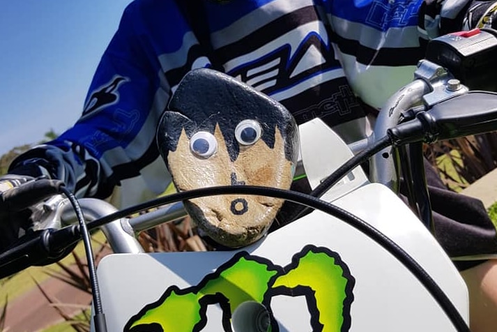 A pet rock sitting on the front of a motocross bike, with a young rider behind.