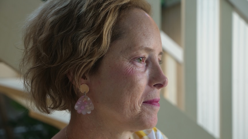 A close-up of a middle-aged woman's profile, short blonde hair wearing pink lipstick, gold and pink earings, looking pensive.