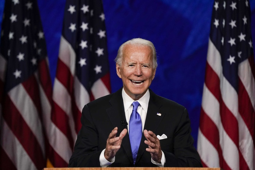 Joe Biden stands at a podium making a speech, while gesturing with his hands. Behind him are three US flags.