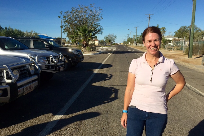 A woman wearing pink t-shirt, blue jeans standing on a road, blue sky and smiling at the camera.