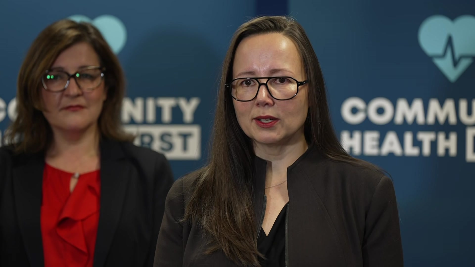 A woman with long dark hair, dark rimmed glasses and a dark jacket stands in front of a blue sign that says "communithy health".