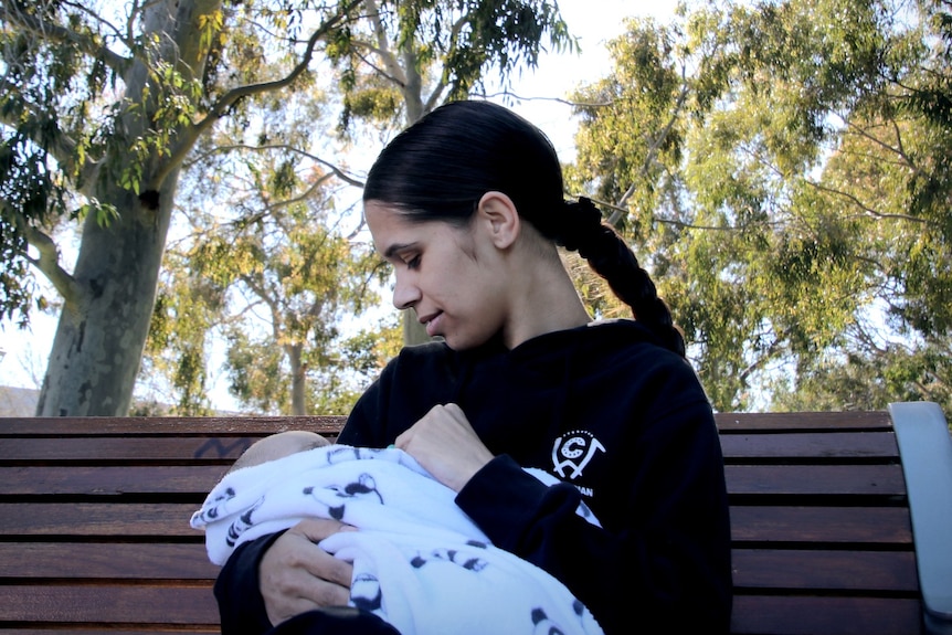 A woman sits on a park bench, looking down at her baby.
