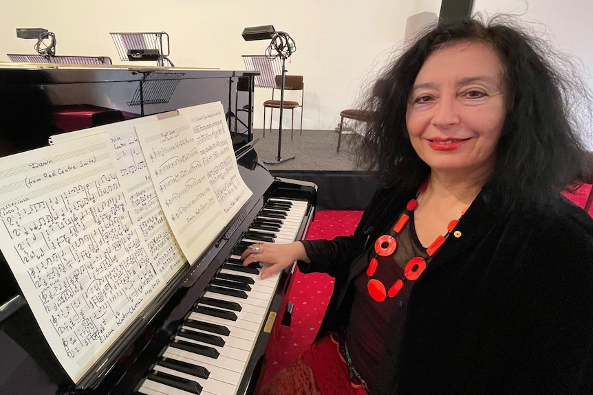 Woman wearing red necklace on black clothing sitting in front of upright piano