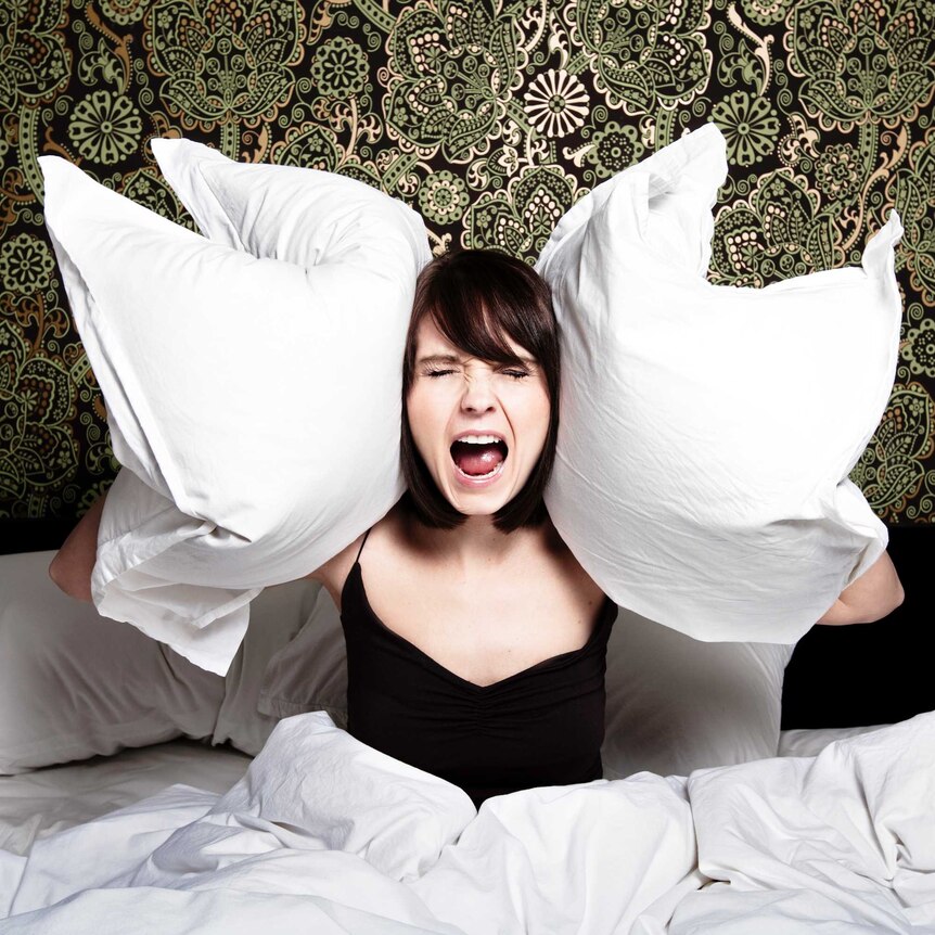 Woman sitting upright in bed with two pillows held to her ears and screaming against a backdrop of green paisley wallpaper