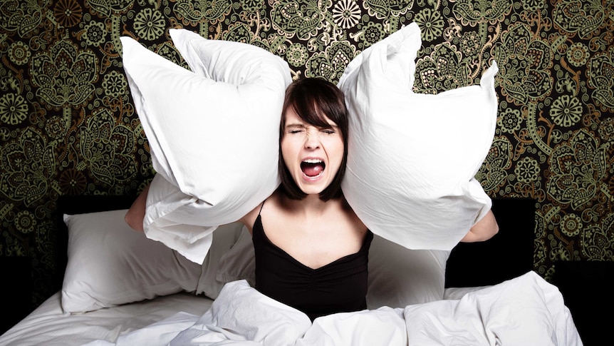 Woman sitting upright in bed with two pillows held to her ears and screaming against a backdrop of green paisley wallpaper